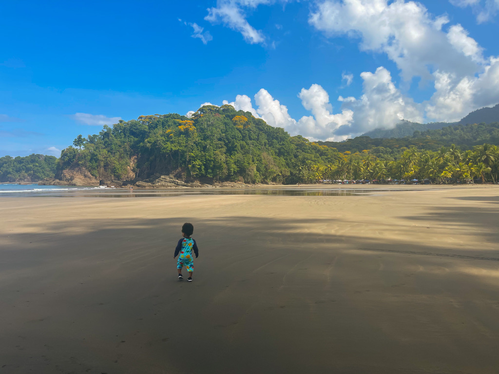 Young child walking on the wide sandy beach at Playa Ventanas with lush green jungle coastline and bright blue sky