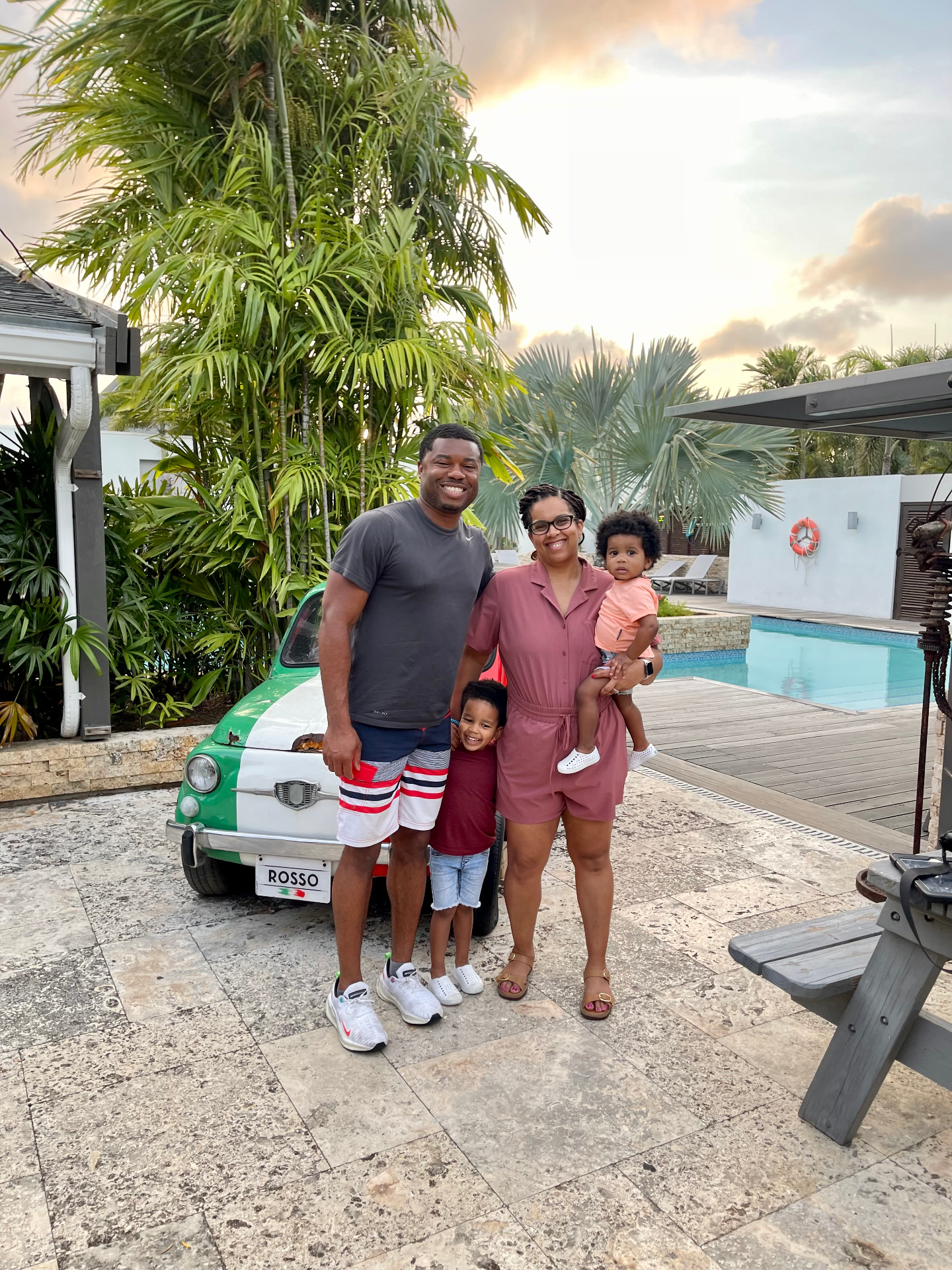 A family of four smiling beside a green and white vintage Fiat at sunset in Curaçao