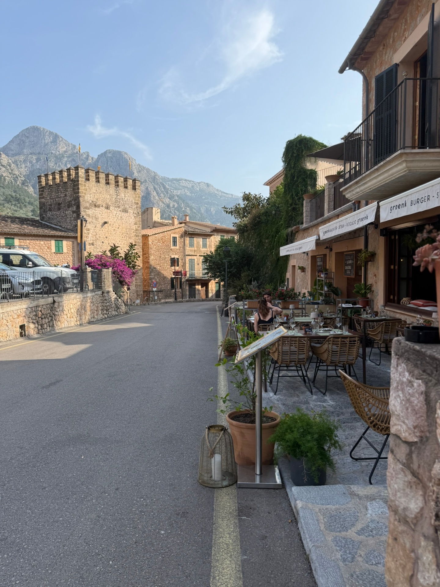 A quiet village street with a stone defense tower and the Tramuntana mountains rising behind cafe terraces