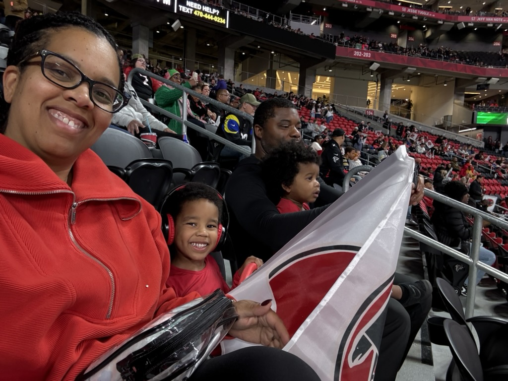 Family in red Falcons gear smiling in the stands at Mercedes-Benz Stadium, one of the boys waving a team flag