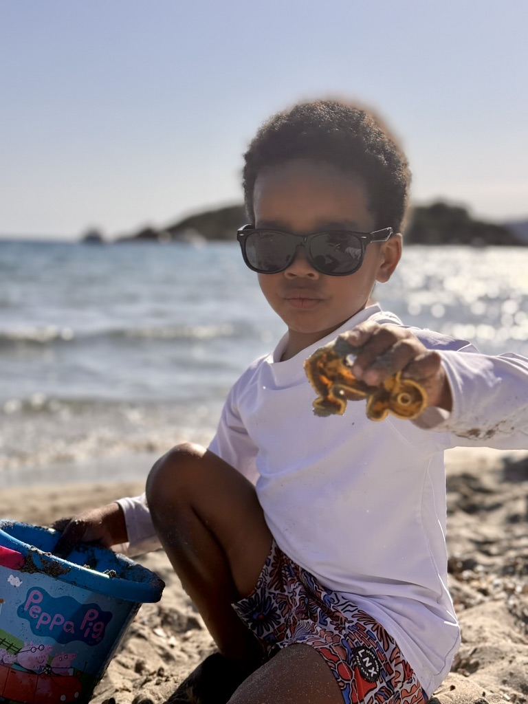 Preschooler in sunglasses holding a sandy toy truck on the beach at Sant Elm