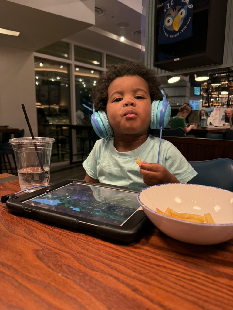 Avery in headphones eating fries at a restaurant table with a tablet propped in front of him