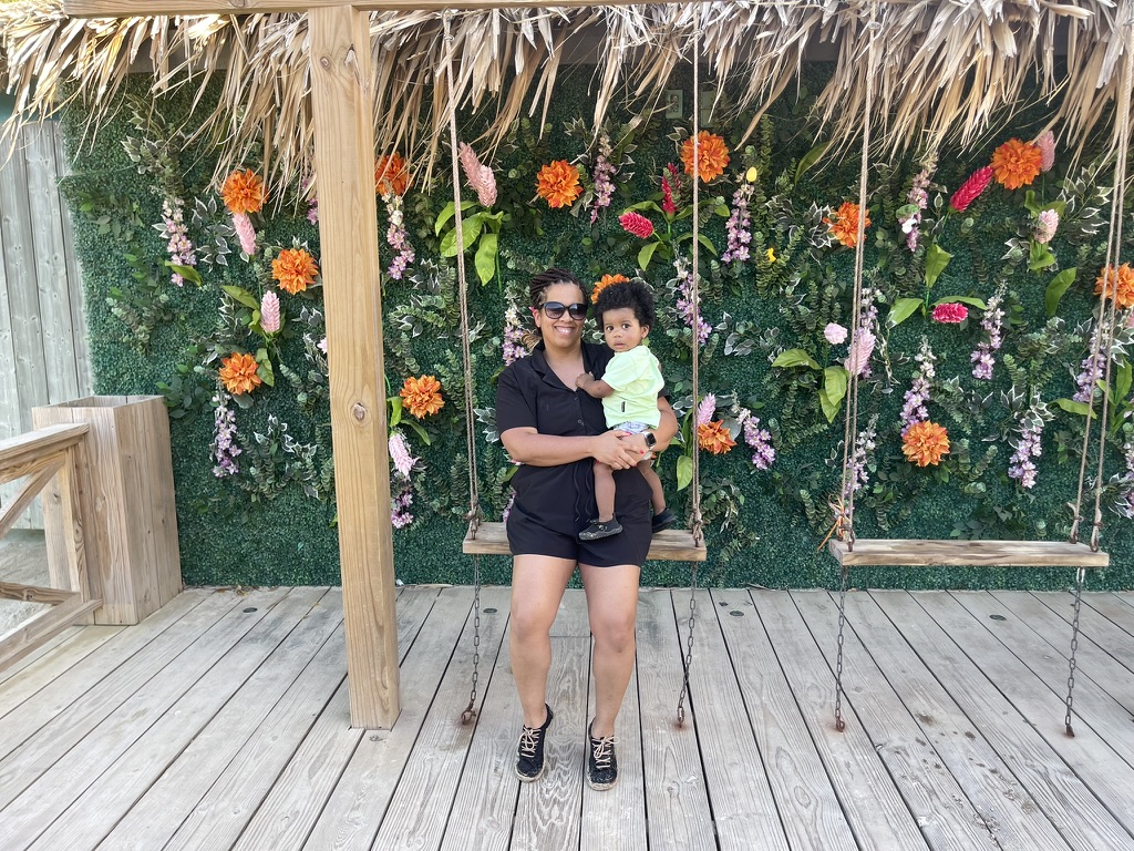 Mother holding her toddler on a beachside swing in front of a flower wall
