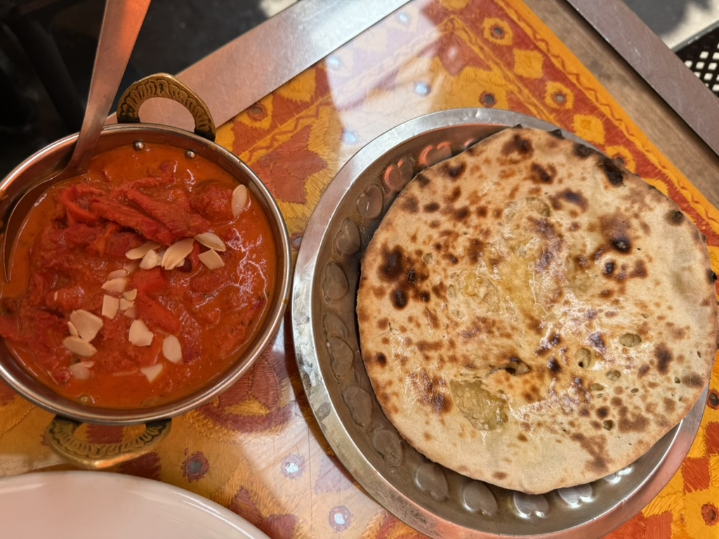 A copper bowl of butter chicken topped with sliced almonds beside fresh naan on a metal plate.