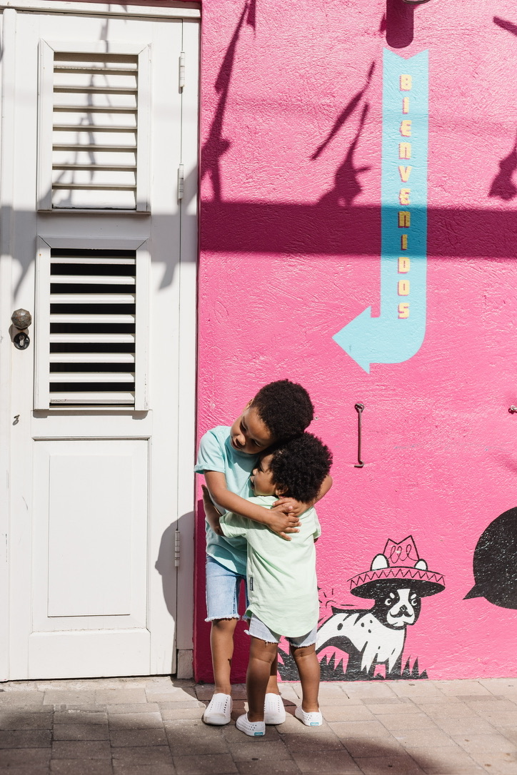 Older brother hugging his younger brother in front of a pink wall painted with a Bienvenidos mural