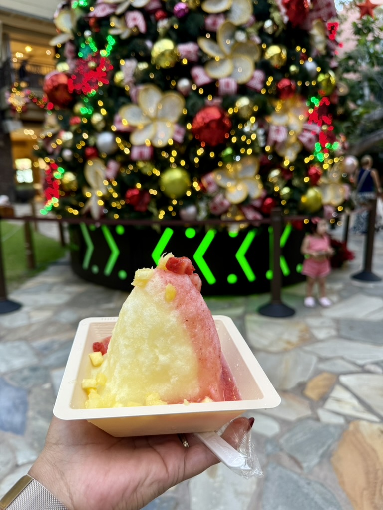 A hand holding strawberry and pineapple shave ice in front of a large decorated holiday tree in Waikīkī