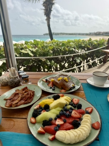 Breakfast of french toast, bacon and a fruit plate on a beachfront table