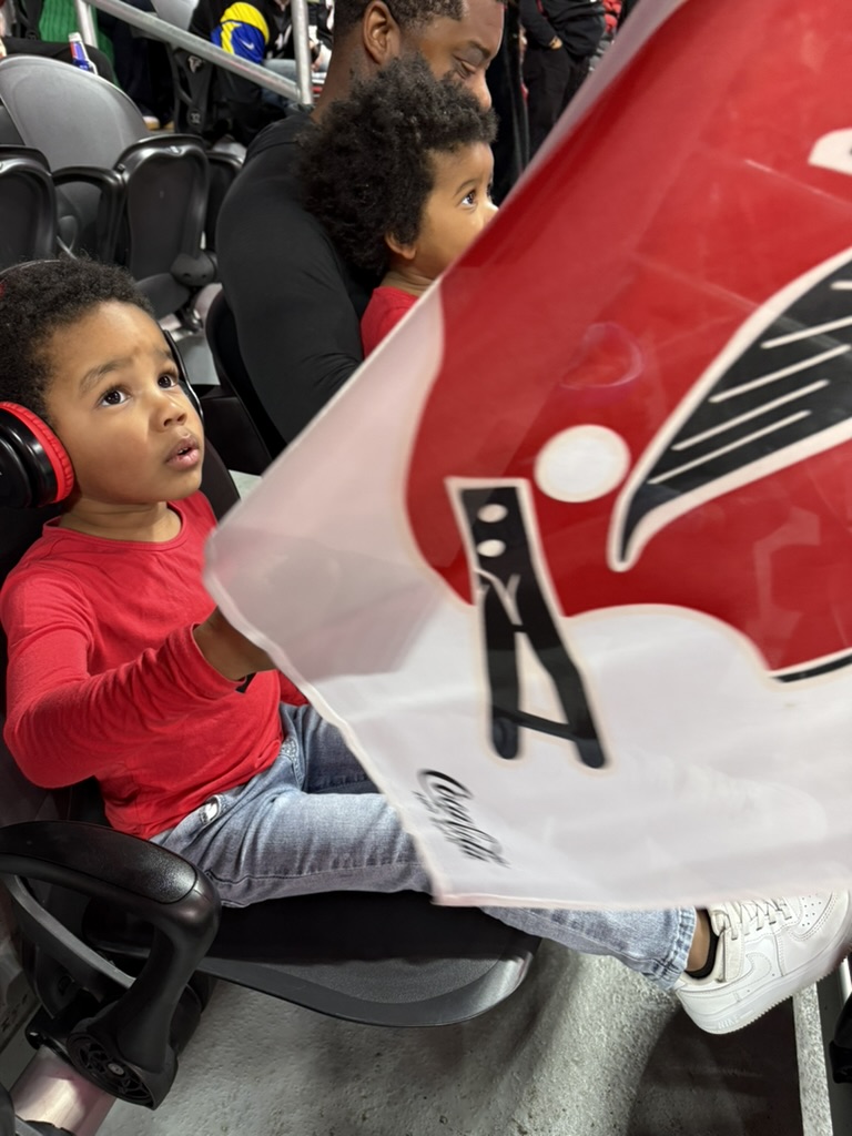 Toddler in red ear defenders sitting in a stadium seat beside his brother and dad, a Falcons flag waving in front of them