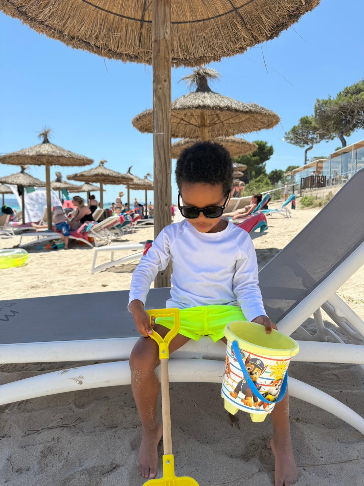 A young child in sunglasses and a white rash guard sitting on a beach lounger under thatched umbrellas, holding a yellow sand bucket and shovel