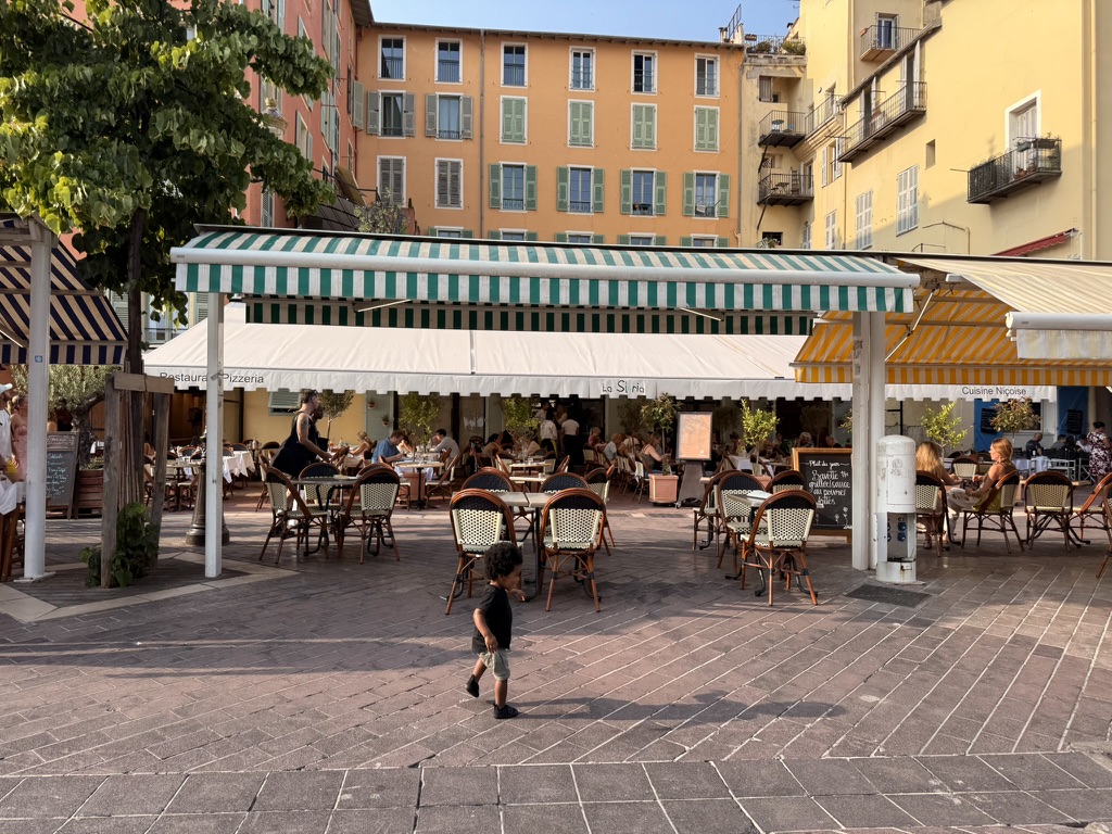 A toddler walking across a sunlit square in Old Nice past striped restaurant awnings
