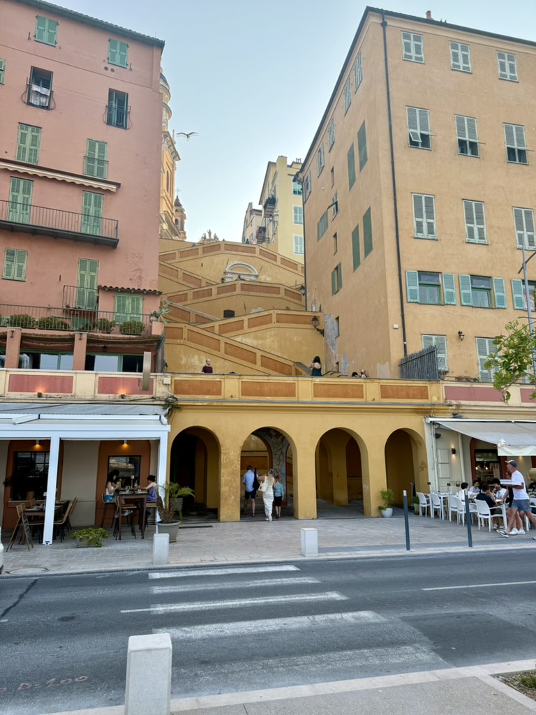 Pink and yellow buildings with green shutters and stone staircases above arcaded cafés in Menton's old town