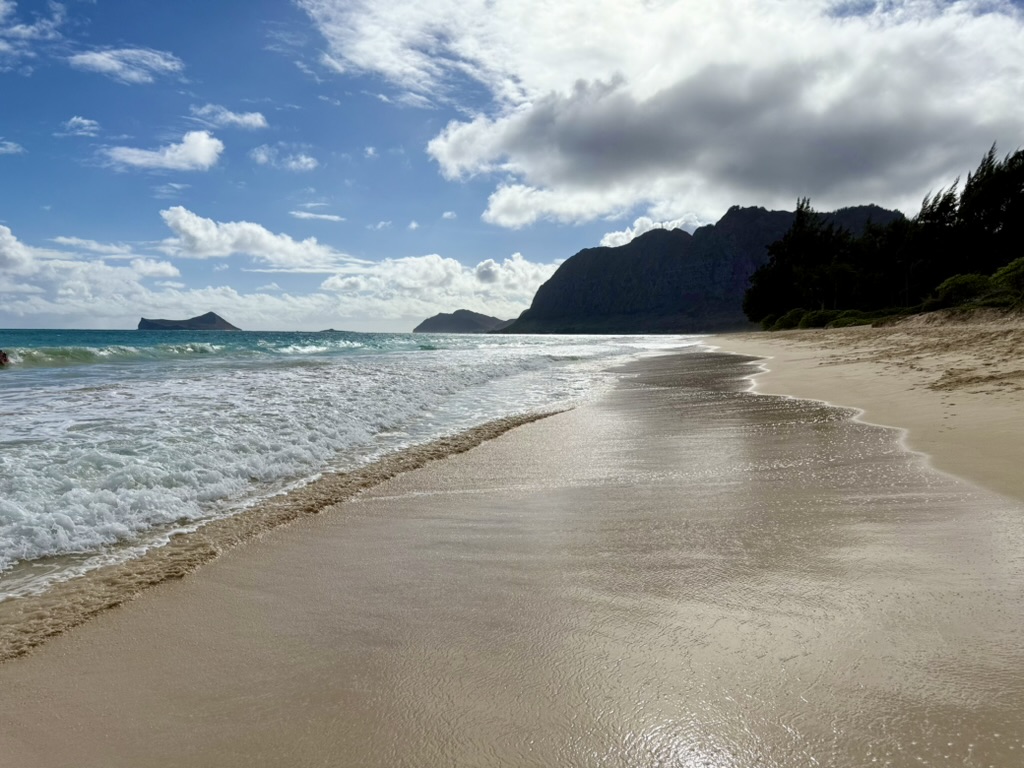 Waimānalo Beach shoreline with turquoise water, white sand and green mountain cliffs under a partly cloudy sky