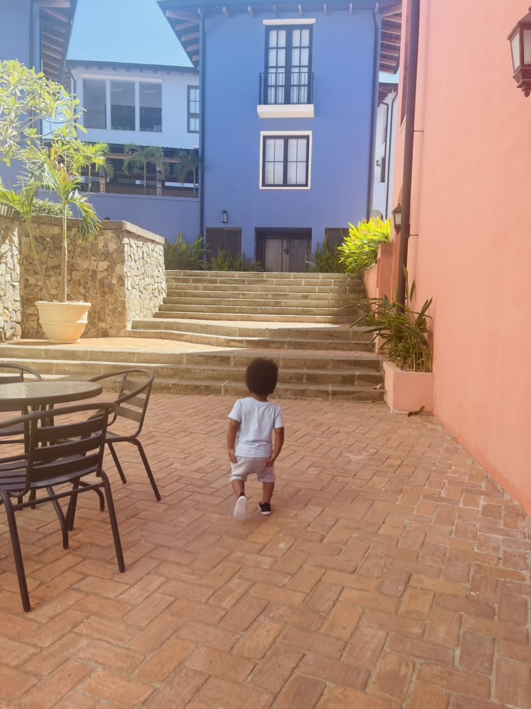 A toddler walking across a sunlit brick courtyard toward colorful buildings and stone stairs in Las Catalinas, Costa Rica