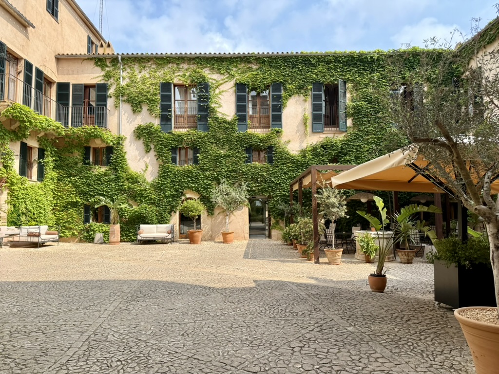 The ivy-covered stone courtyard of the Zoëtry estate with green shutters, potted olive trees, sofas and a shaded terrace