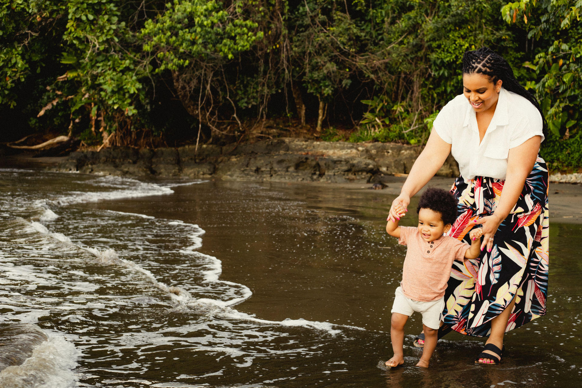A mother holds her toddler's hands as they stand together at the edge of the ocean