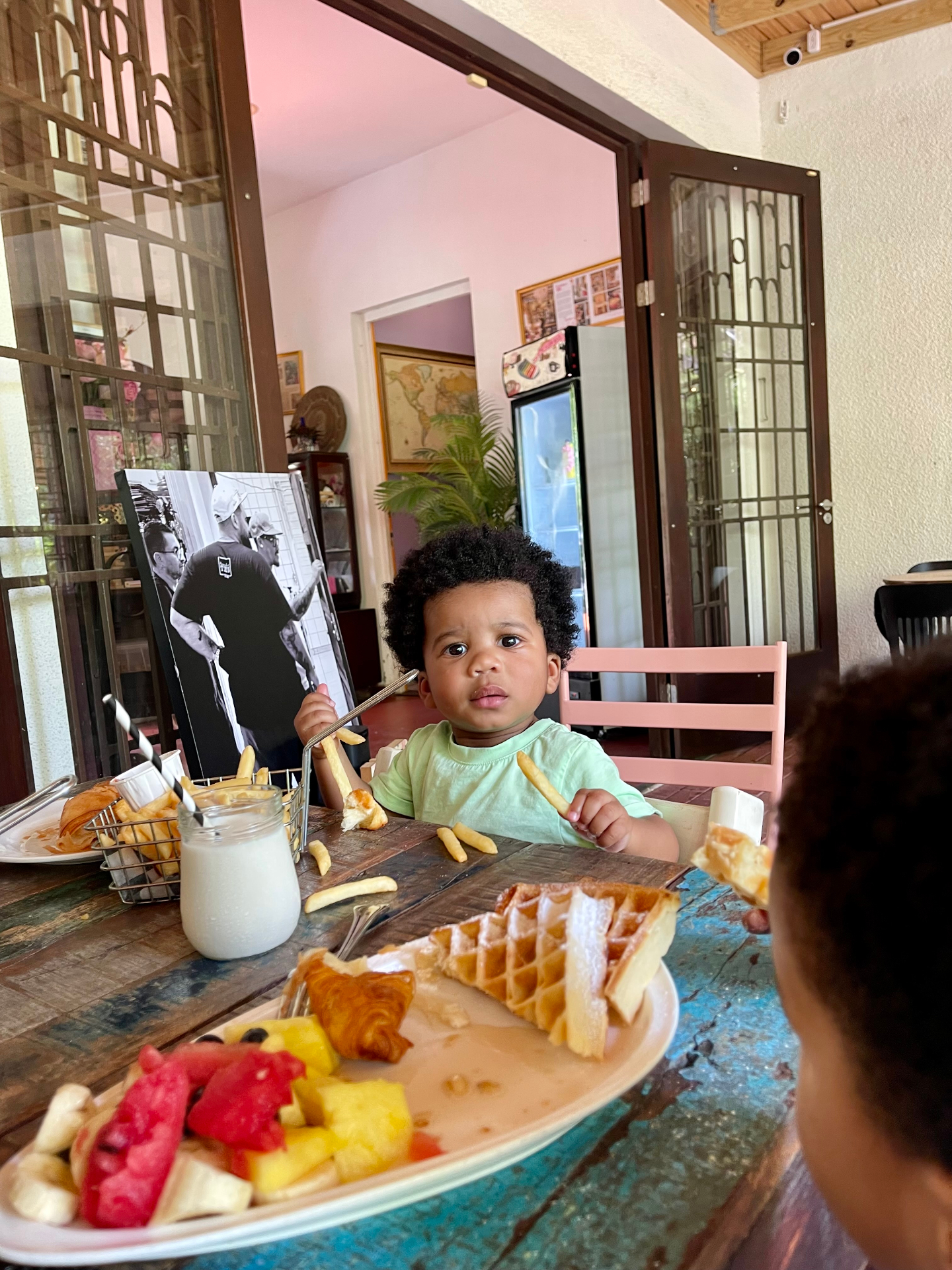 A toddler at a colorful restaurant table with waffles, fruit and fries
