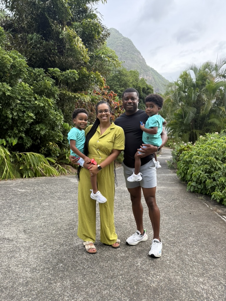 A smiling family of four standing together on a tropical path with lush greenery and mountains behind them