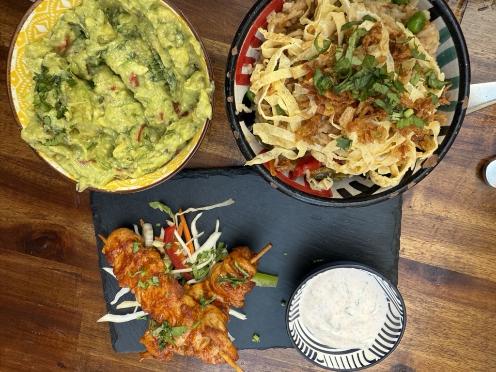 Overhead table shot at Hacienda with a bowl of rice topped with crisp tortilla strips, a bowl of guacamole, a slate board of chicken skewers and a bowl of white sauce