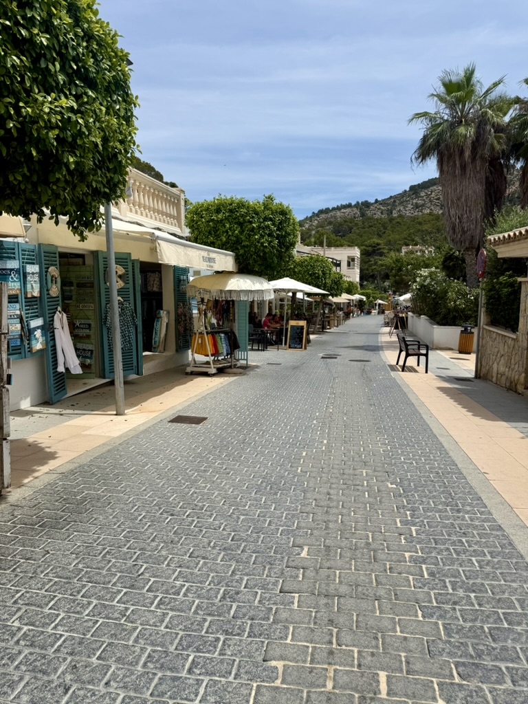 A quiet cobblestone pedestrian street in Sant Elm lined with shuttered shops, cafe umbrellas and palms, with pine-covered hills behind