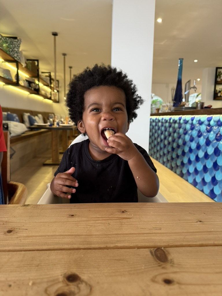 Toddler in a high chair grinning mid-bite at a waterfront restaurant in Sant Elm
