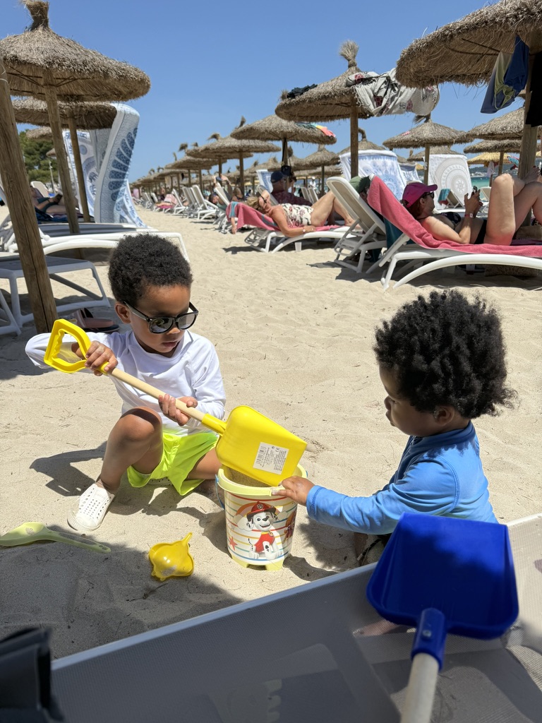 Two young brothers filling a sand bucket with shovels among rows of thatched umbrellas and loungers at Playa de Muro