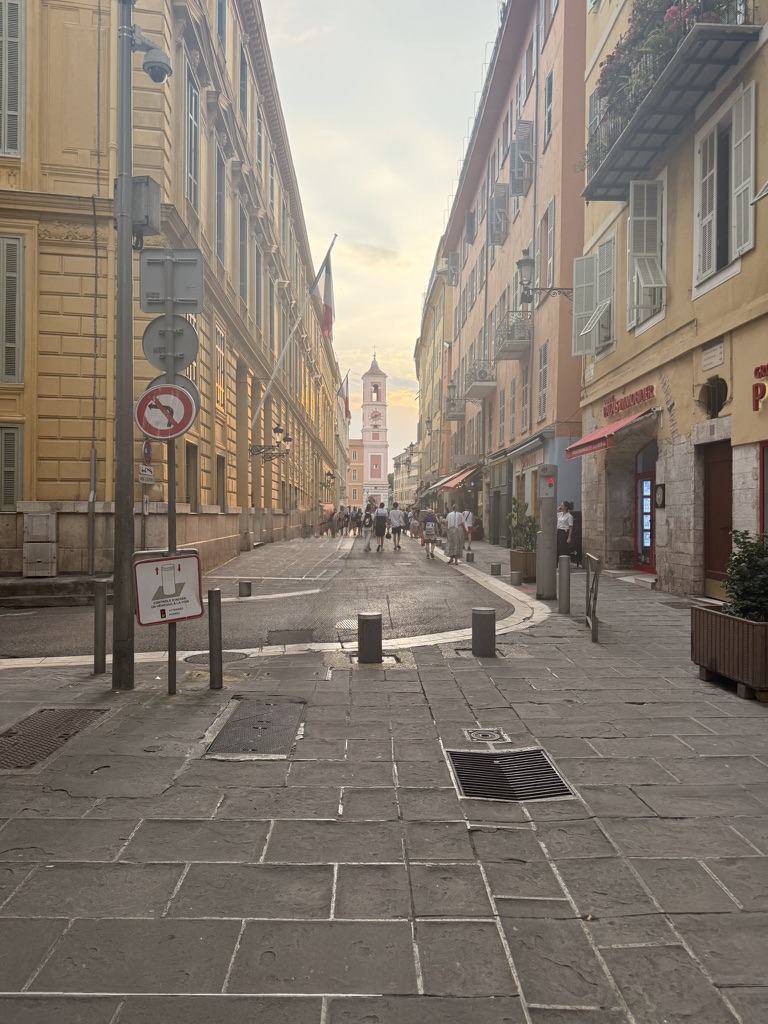 A pedestrian street in Old Nice at golden hour with pastel buildings and a bell tower in the distance.
