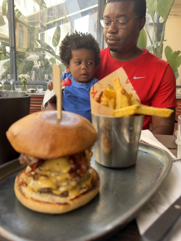 A father holding his young son at a restaurant table with a tall burger and a tin of fries in the foreground.