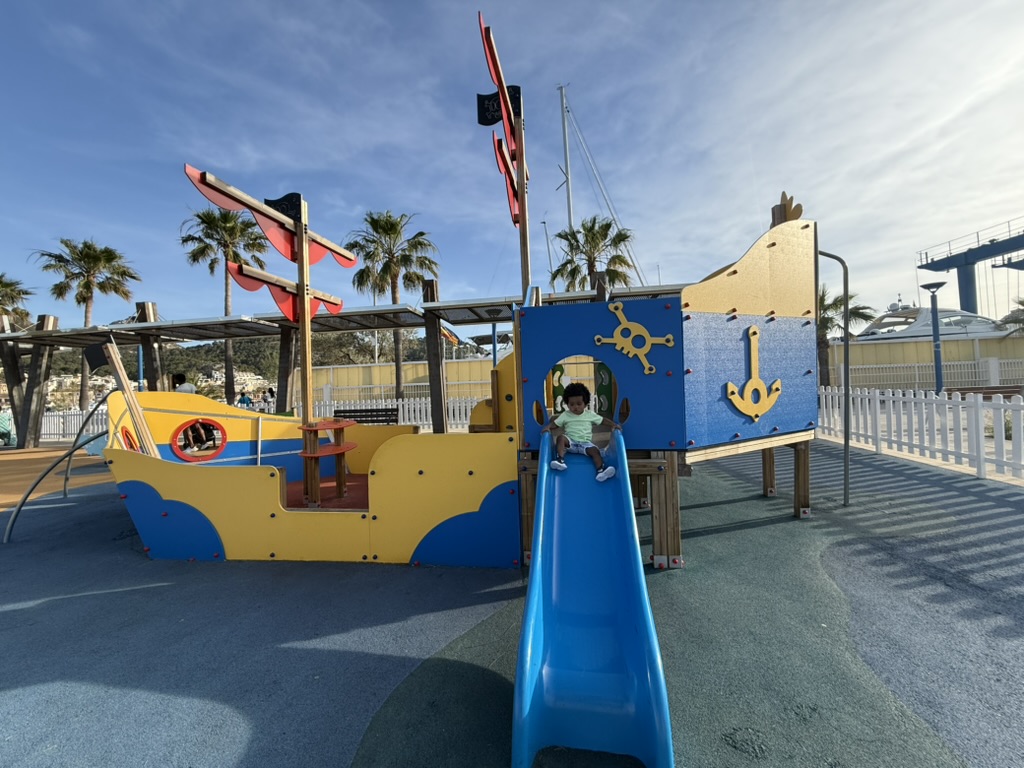 Child coming down the blue slide of the yellow pirate ship playground at Port d'Andratx