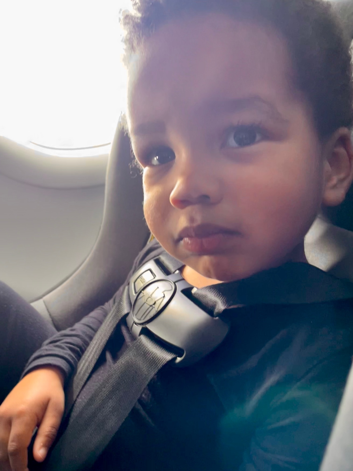 A child sitting in an airplane seat wearing a harness and looking toward the camera