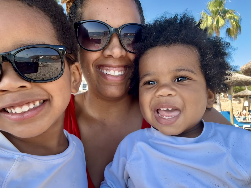 A mother smiling on a sunny beach with two young children in rash guards, palm trees and thatched umbrellas behind them