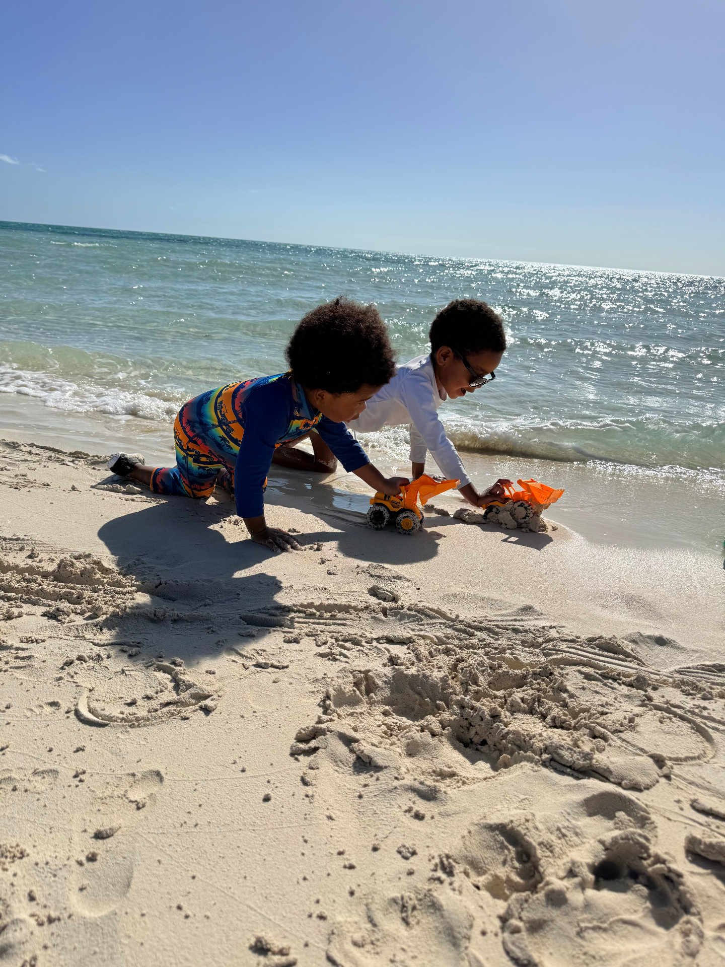Two children playing with toy trucks on a sandy beach at the water's edge