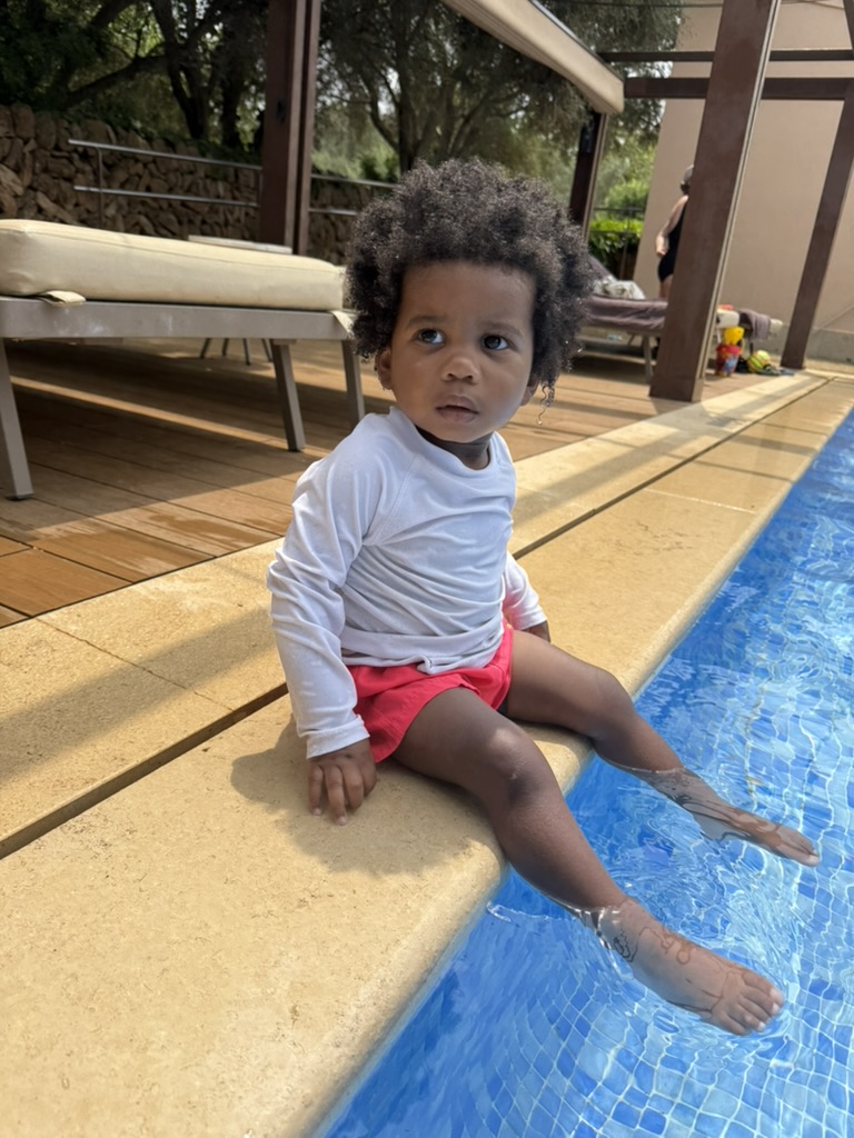 A toddler in a white rash guard sitting on the edge of the pool with feet in the water