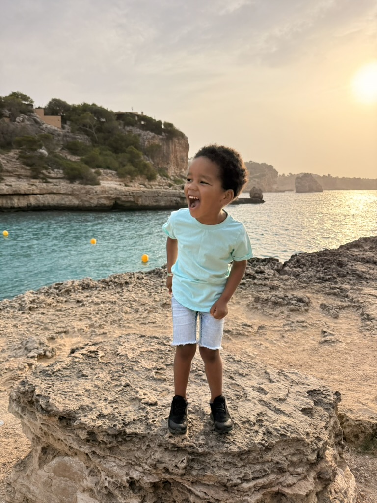 A young child in a light blue shirt standing on rocky cliffs above the turquoise water of Cala Llombards at golden hour