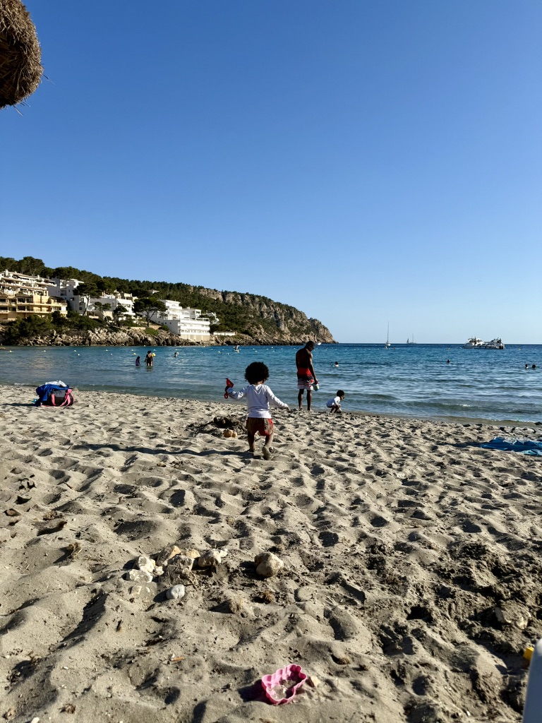 A toddler running toward the calm turquoise water at Sant Elm beach, with white village buildings and a pine-covered headland behind