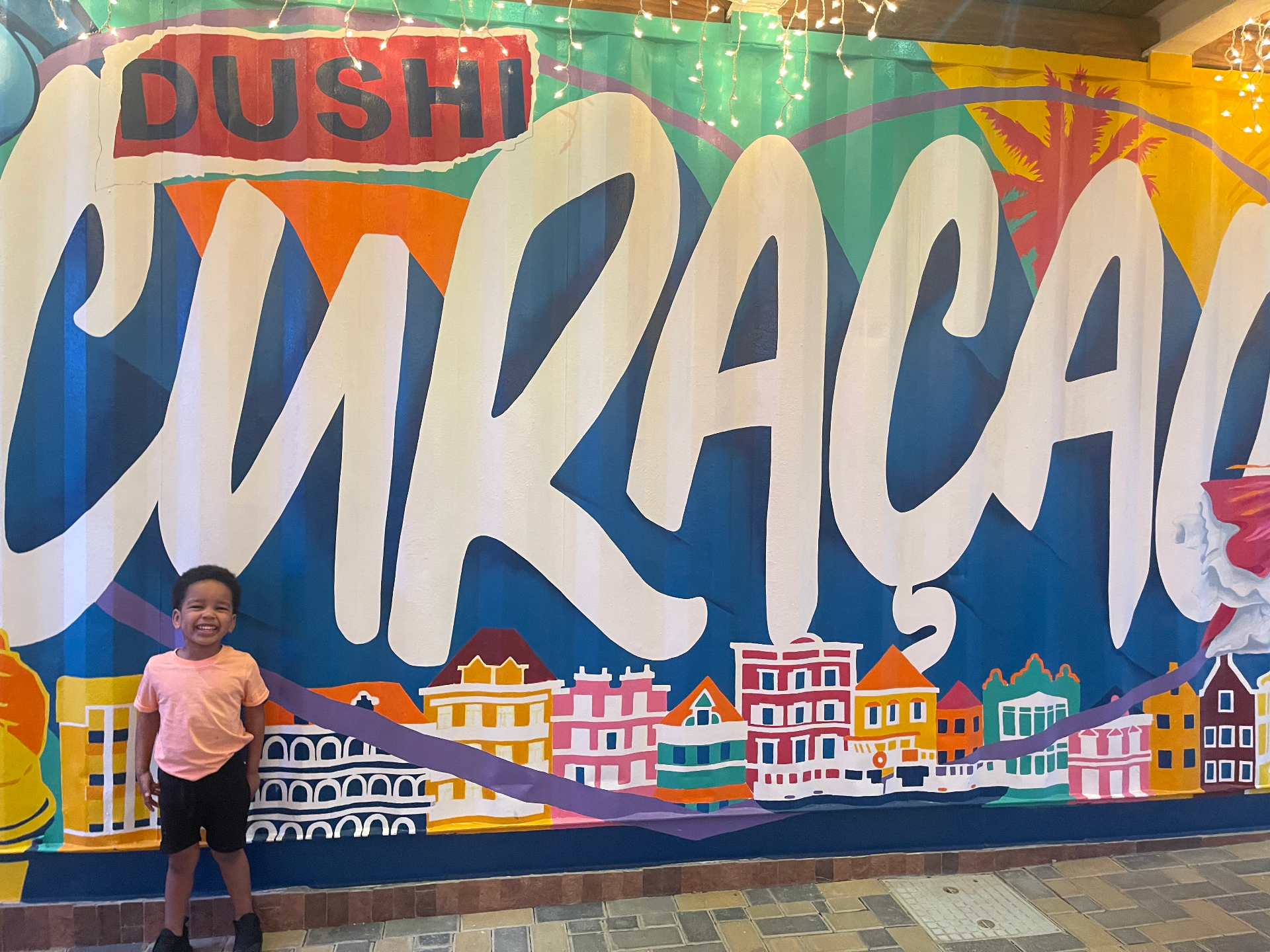 A young boy standing in front of a bright Dushi Curaçao mural