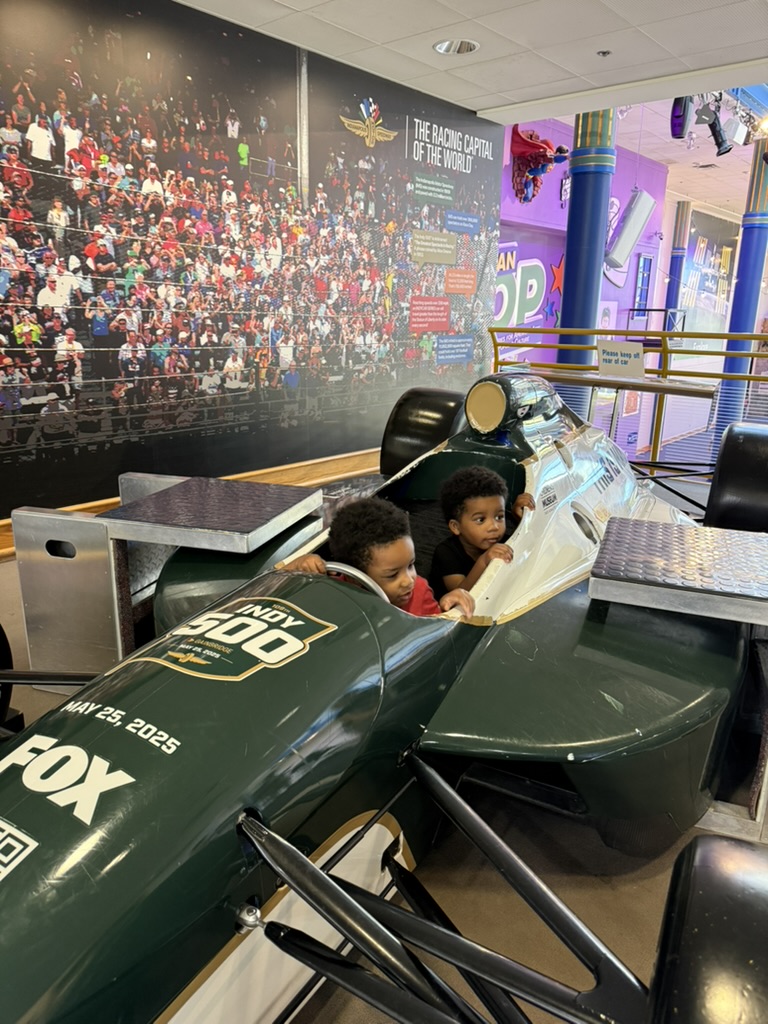 Both boys climbing into the Indy 500 race car inside the museum, with the crowd mural behind them