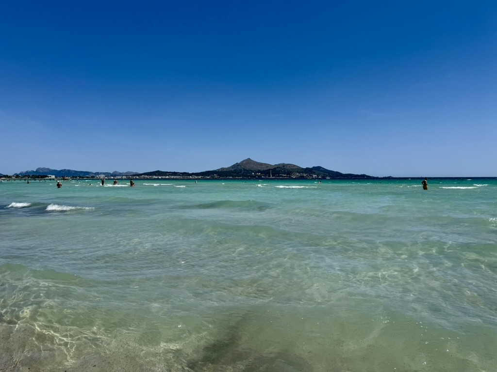 Wide view of the clear turquoise shallows at Playa de Muro with distant headlands under a deep blue sky