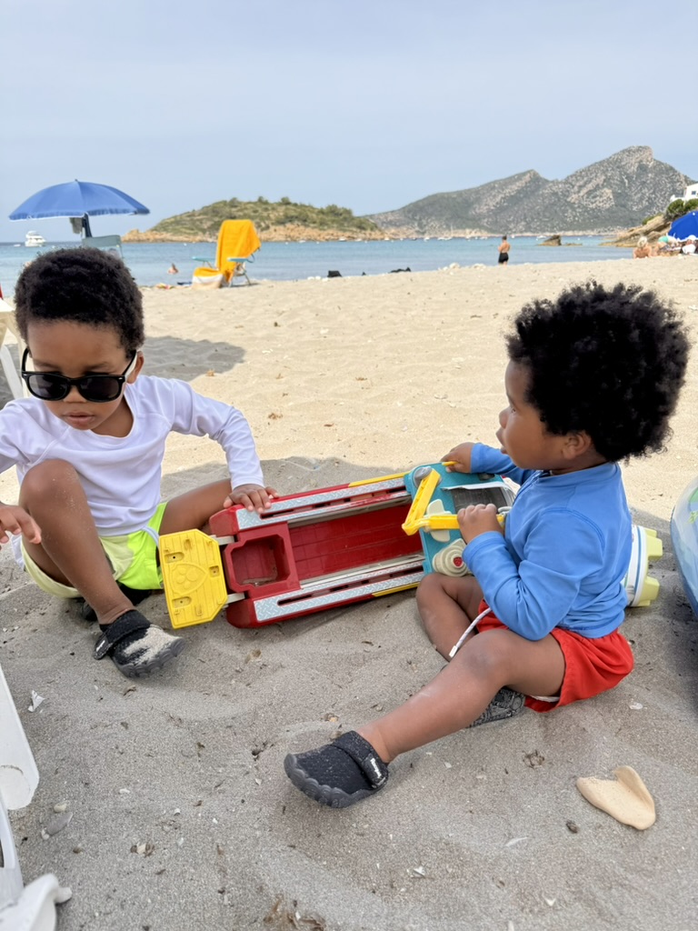 Two brothers playing with a toy fire truck in the sand at Sant Elm with the island headland behind them