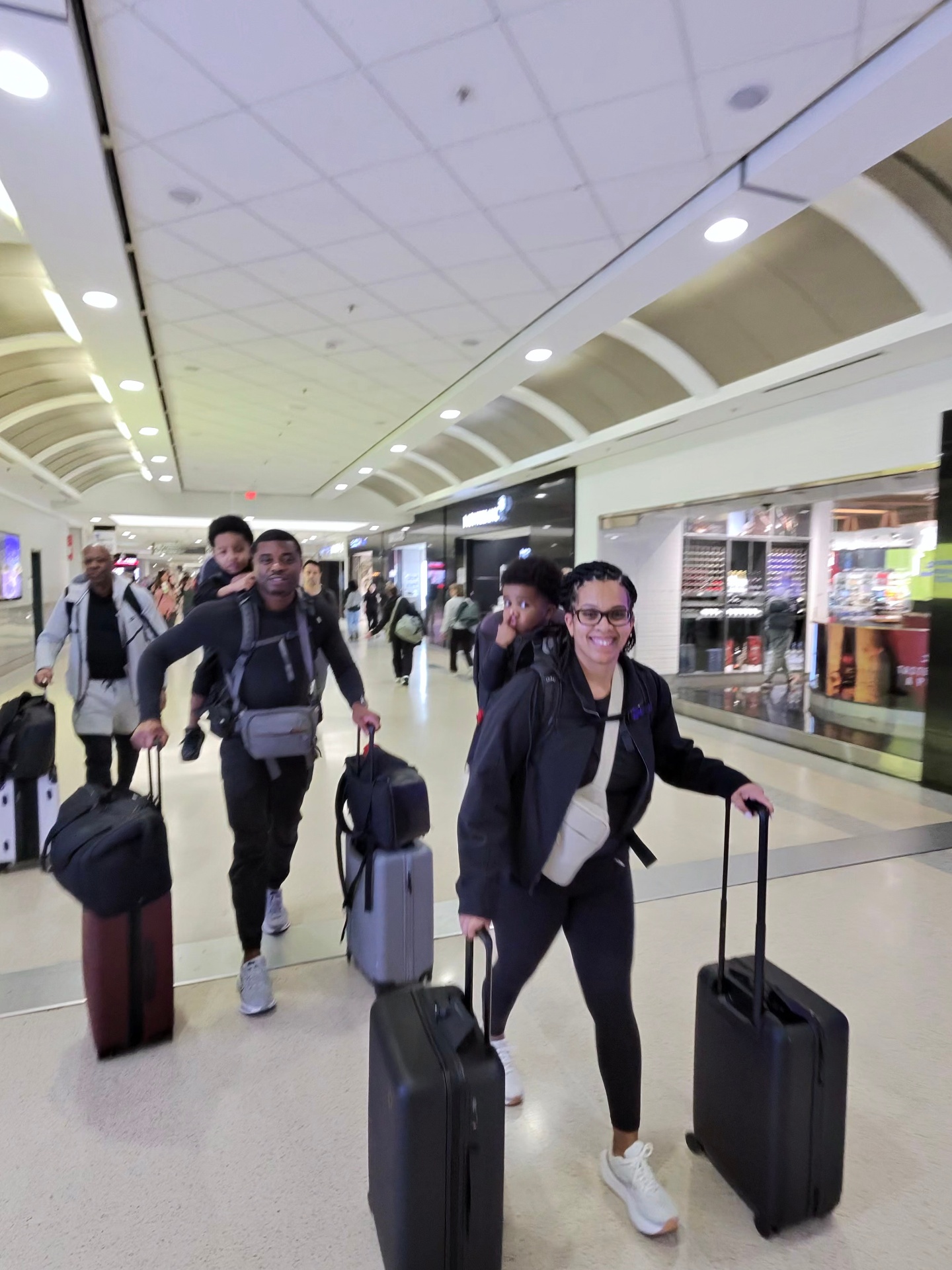 A family walking through an airport terminal with rolling suitcases and children riding on their parents' shoulders