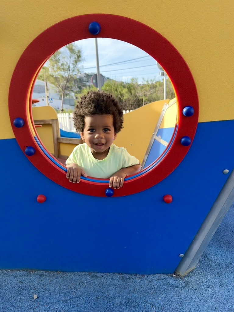 Toddler grinning through the red porthole of a boat-shaped play structure at the port playground
