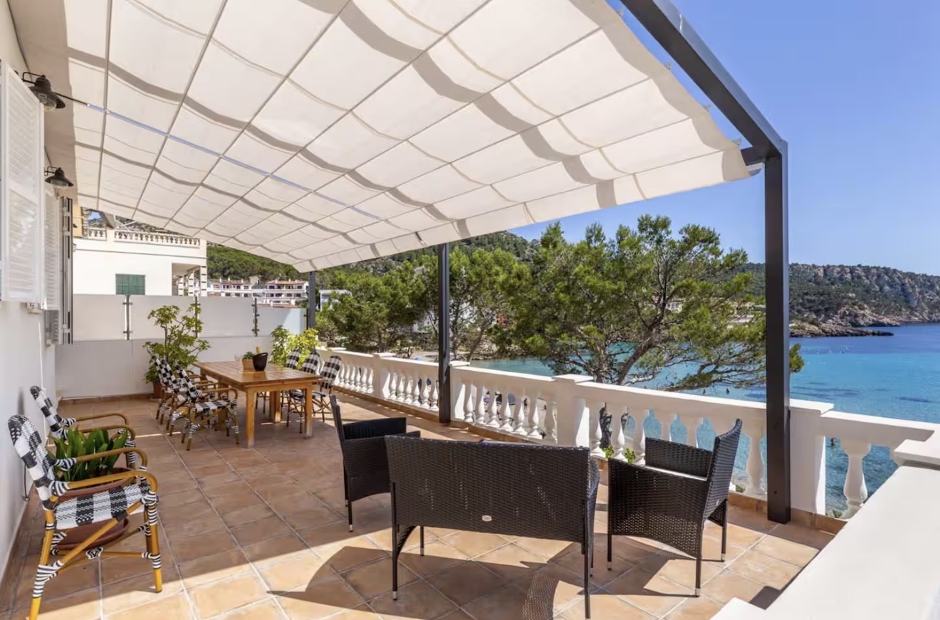 Shaded seaside terrace of the Sant Elm apartment with a long wooden dining table, wicker chairs and an unobstructed view over the turquoise cove