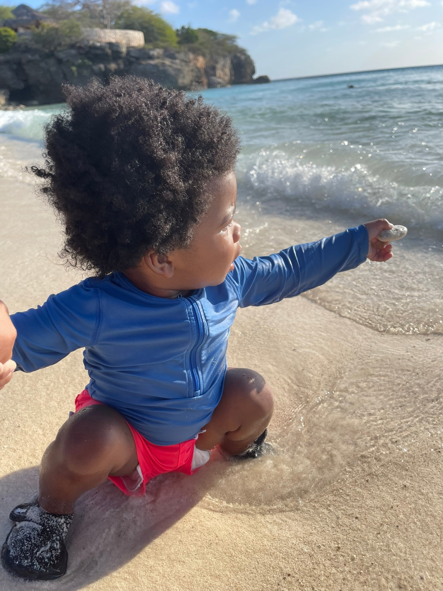 Toddler in a blue rash guard crouching at the shoreline holding a smooth stone