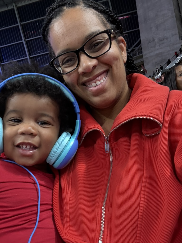 Mother and toddler in red Falcons gear smiling together in the stadium seats, the child wearing light blue headphones