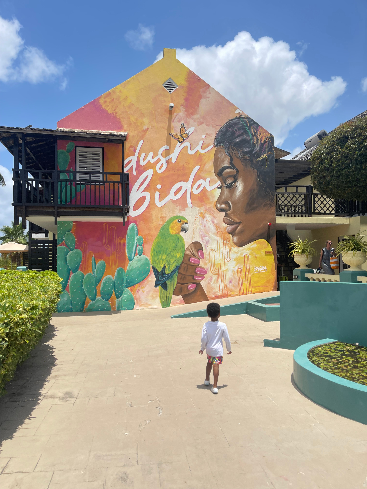 Toddler standing before a bright Dushi Bida mural with a parrot and cacti in Curaçao