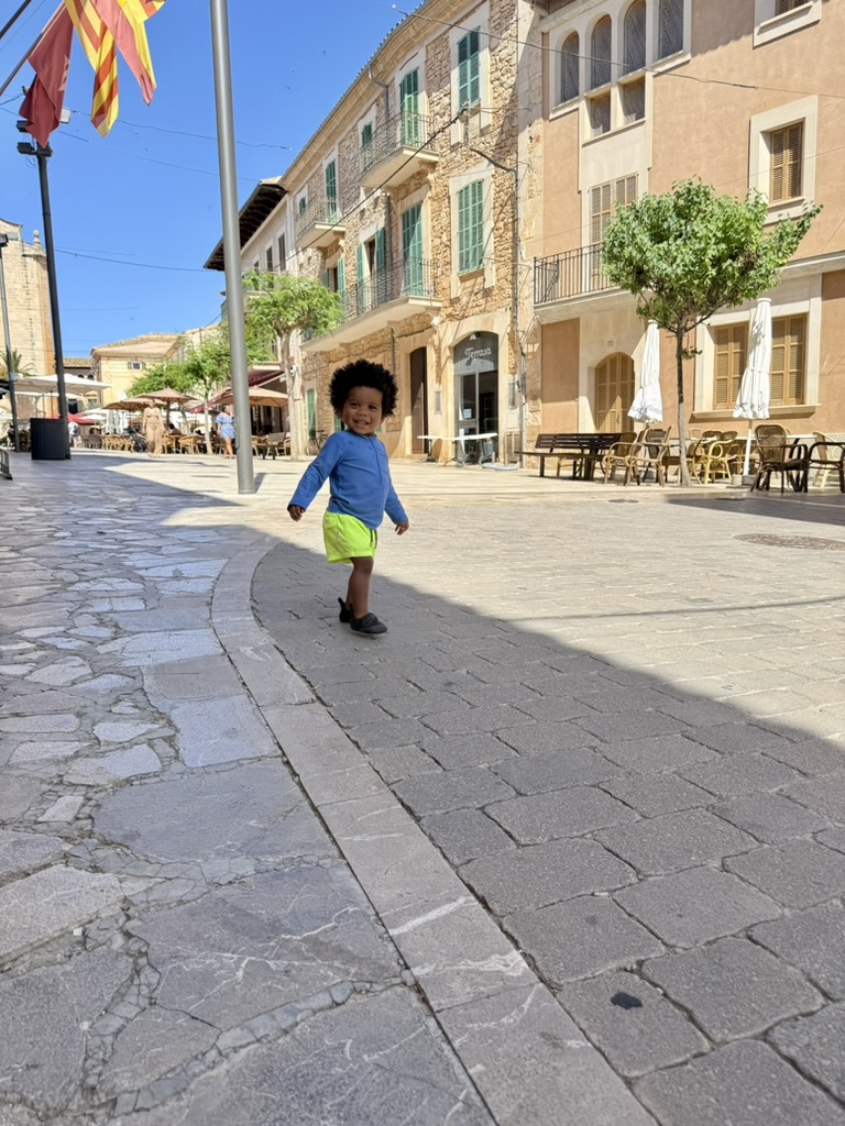 A toddler walking across Placa Major in Santanyi with cafe terraces and flags in the background