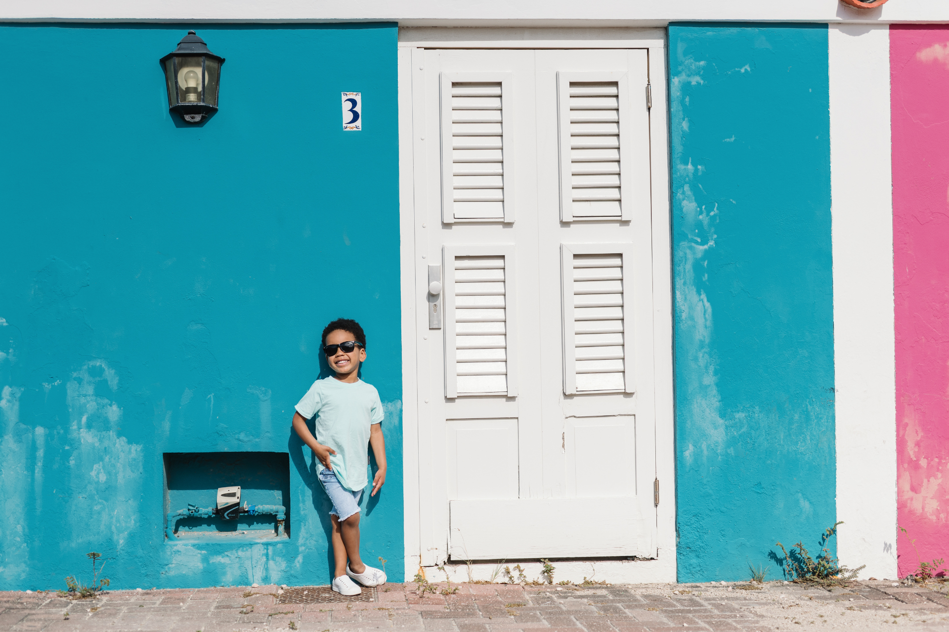 A child leaning against a bright turquoise wall beside a white shuttered door in Pietermaai, Willemstad.