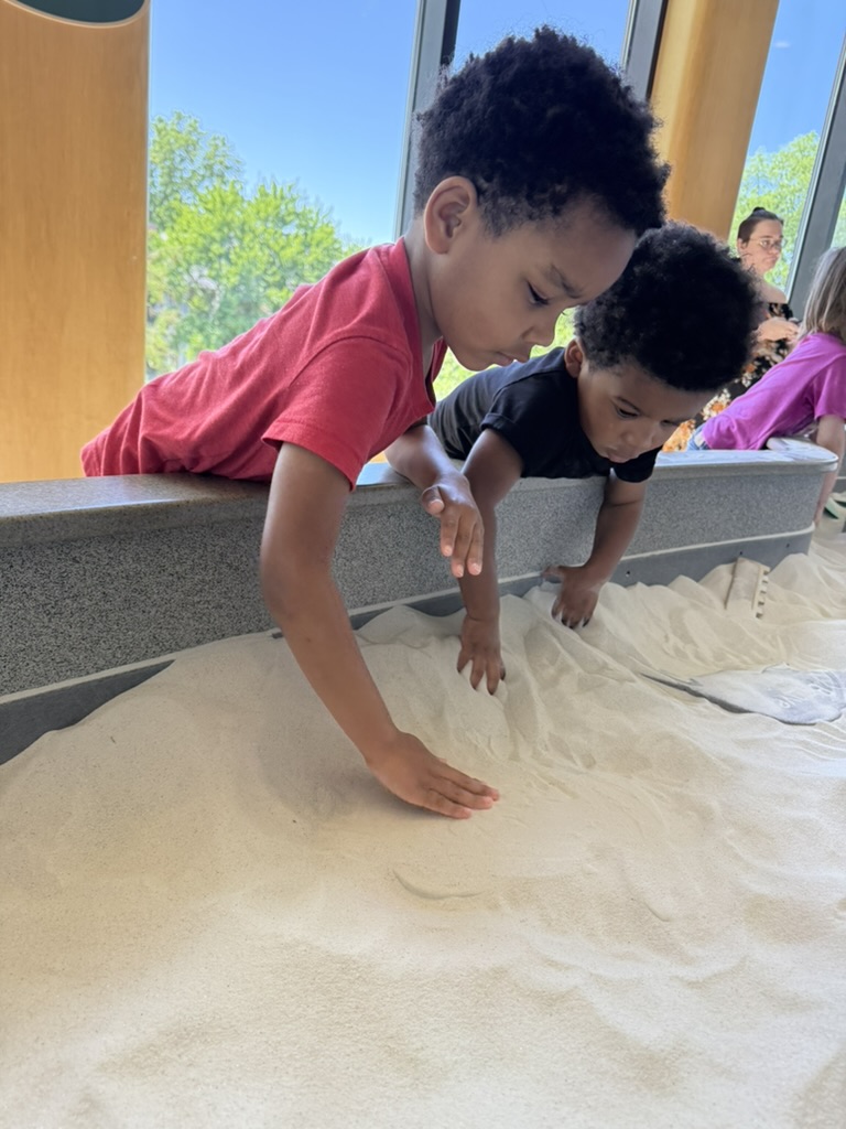 Both boys leaning over the sand table in Playscape, digging with their hands