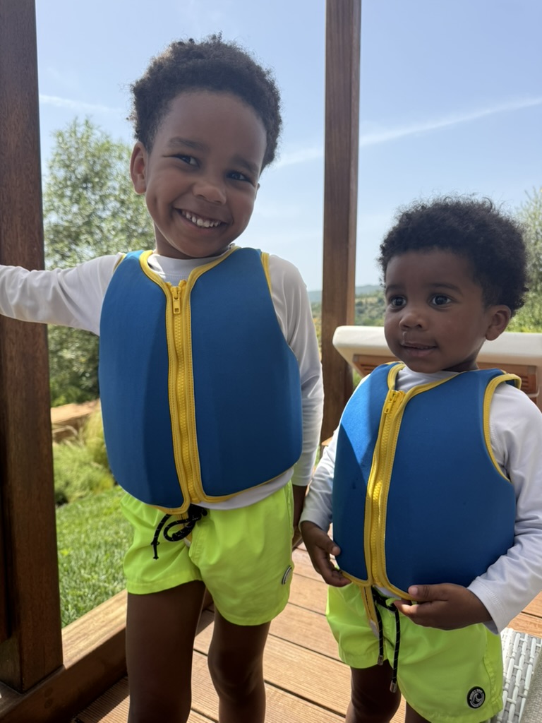 Two young boys in blue and yellow life vests and neon green shorts standing on a wooden terrace with the Algarve countryside in the background
