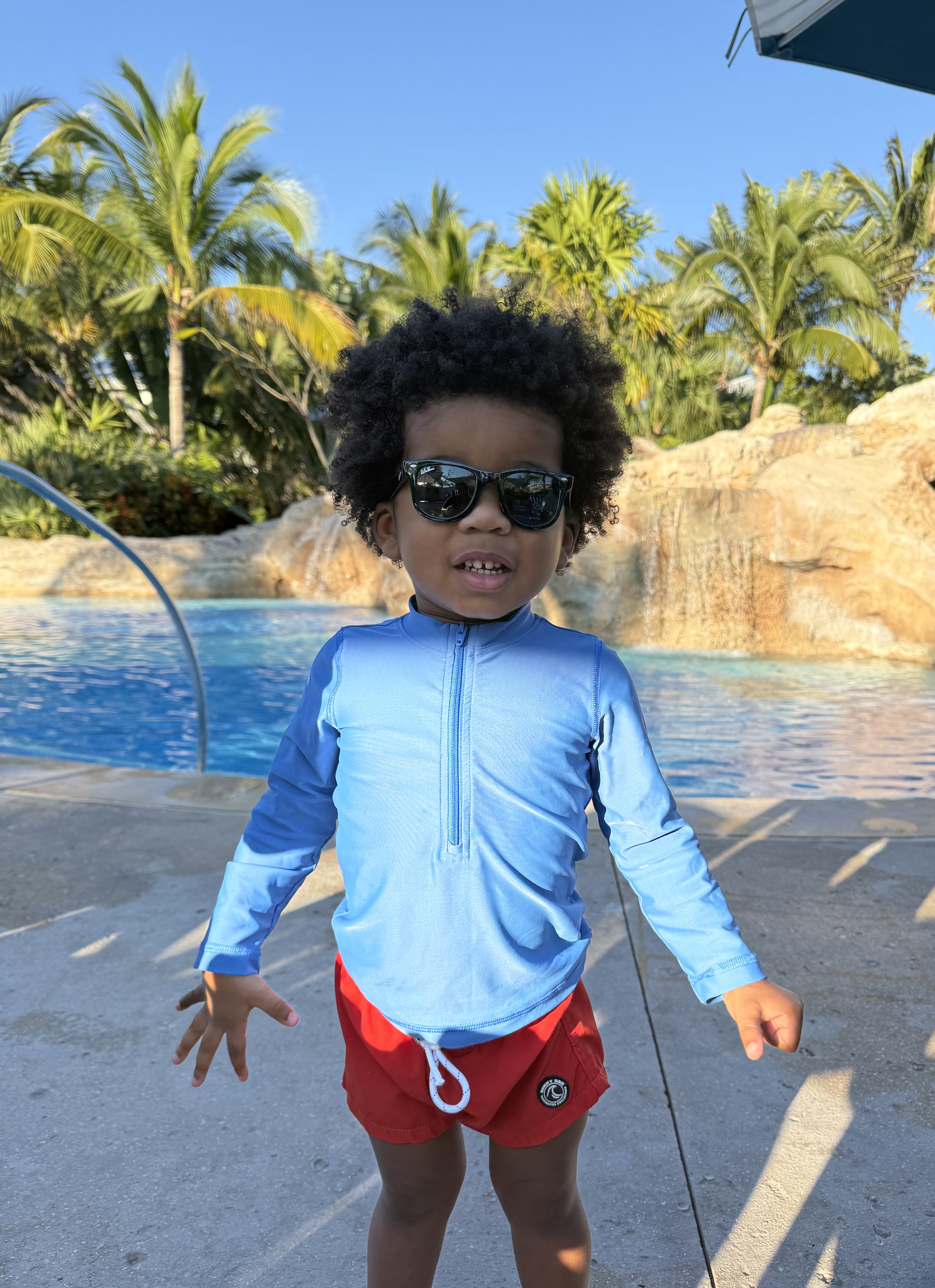 Avery in sunglasses and a rash guard standing poolside with palms and the rock waterfall behind him