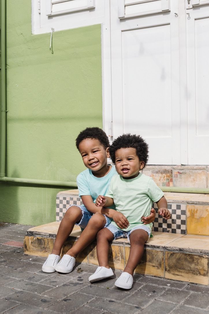 Two brothers in mint shirts sitting together on tiled steps in front of a green Pietermaai house